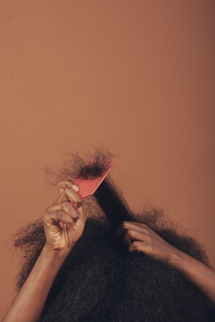 Hands lifting a pick through natural hair against a warm terracotta wall